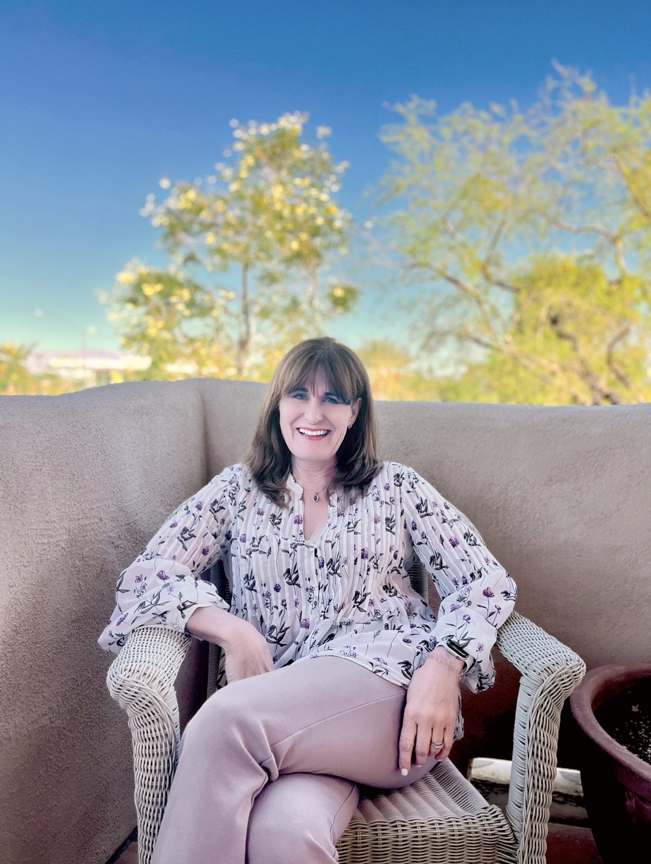 A smiling woman sitting on an outdoor sofa with trees in the background.