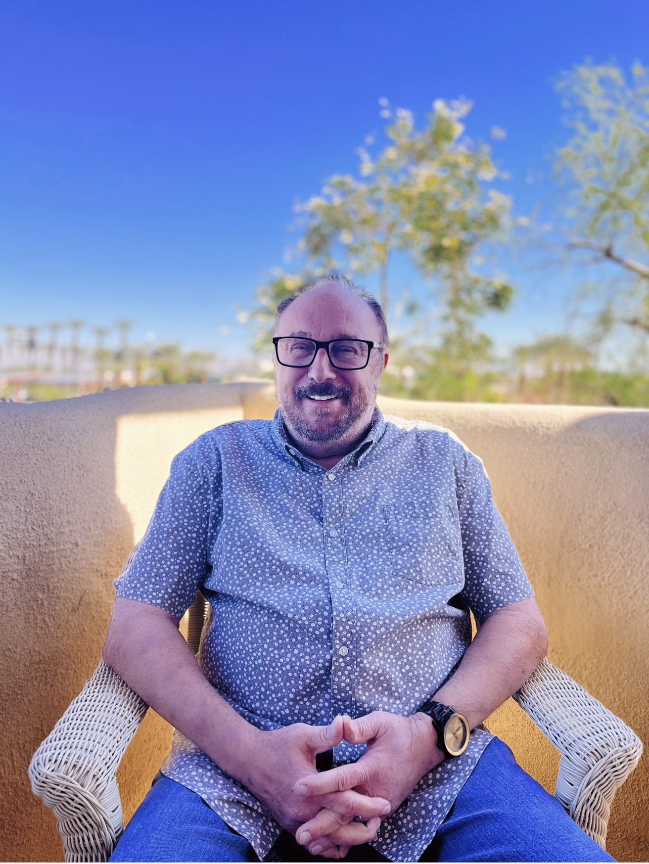 Smiling man with glasses seated outdoors on a sunny day.