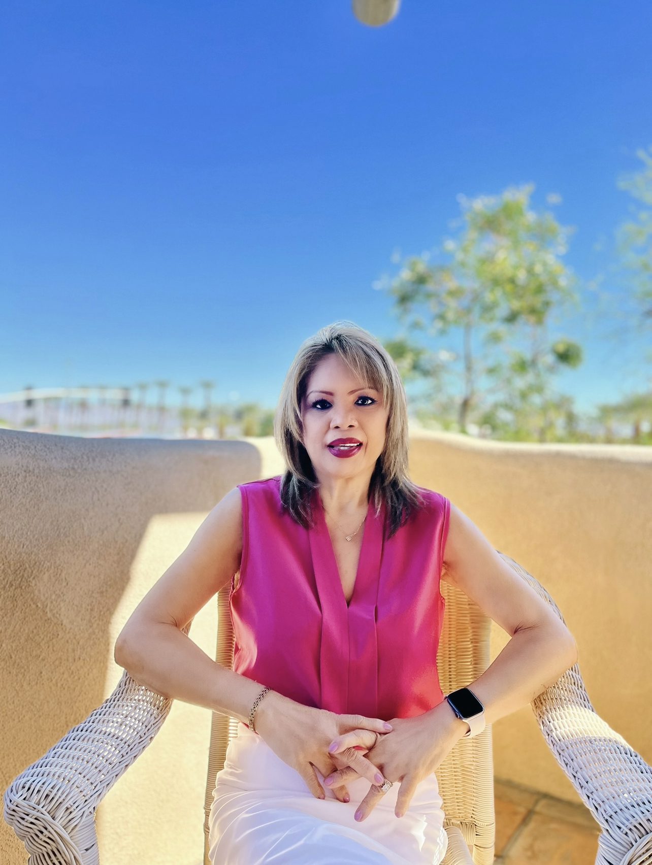 A woman with short hair wearing a sleeveless pink top and white pants sitting on a chair outdoors with a clear sky in the background.