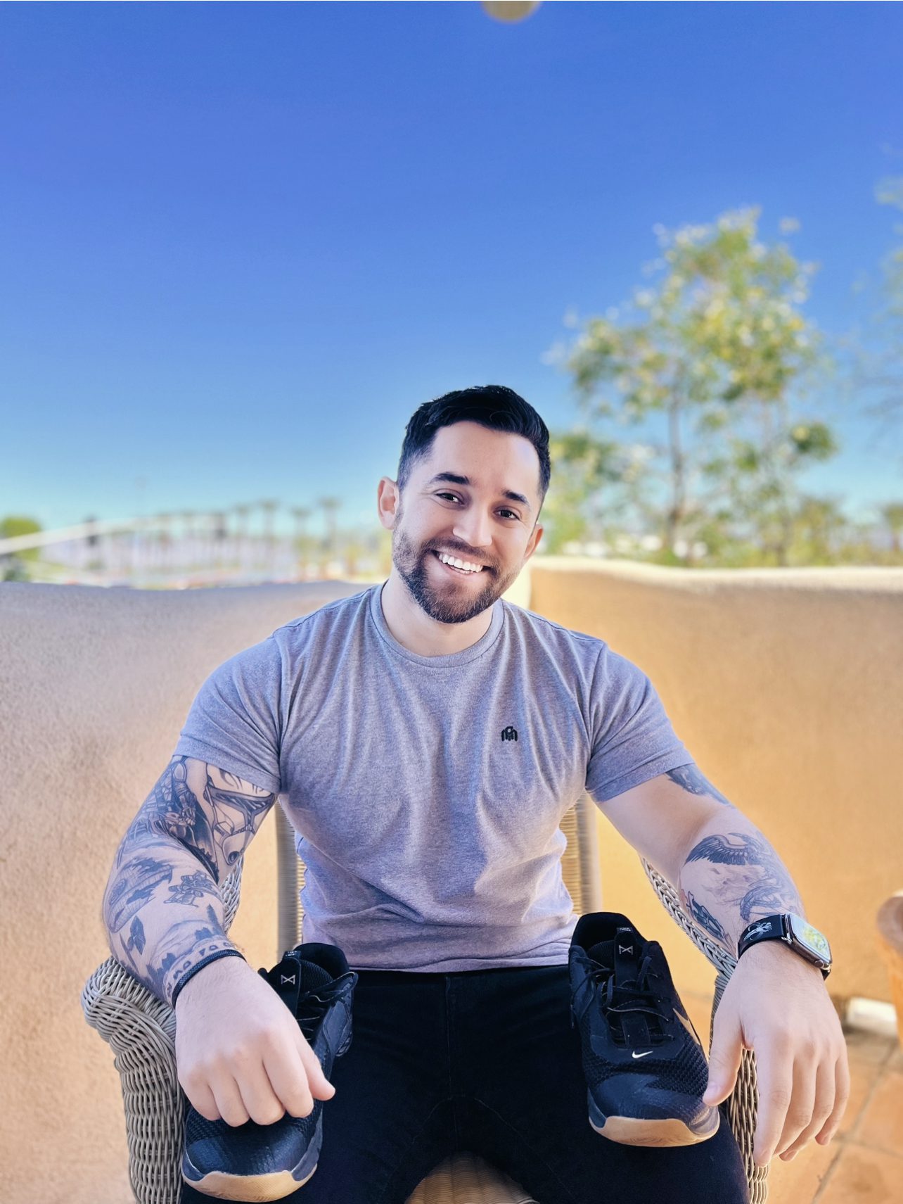 Man with tattoos sitting outdoors, holding a pair of sneakers and smiling at the camera.