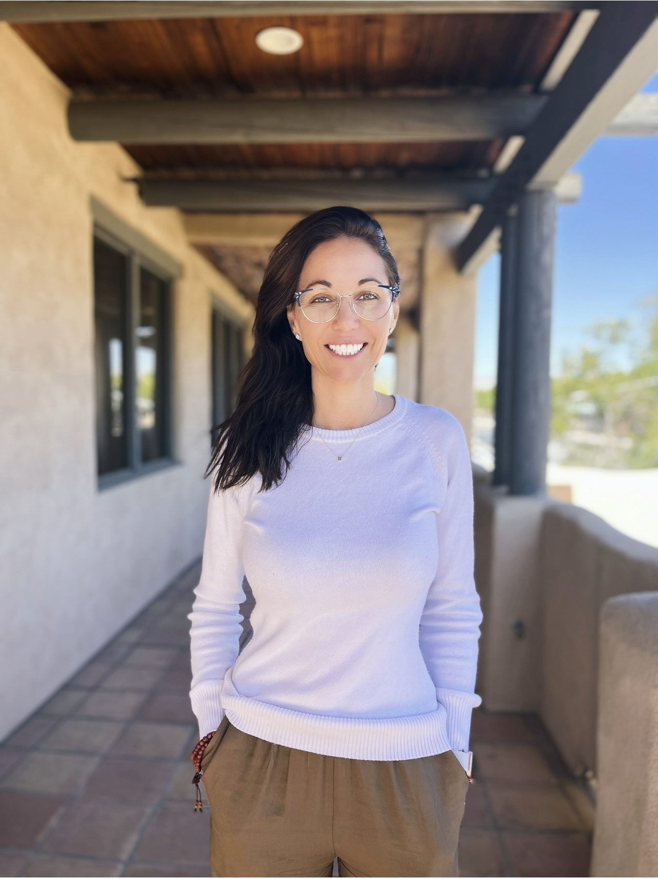 Woman smiling in glasses, wearing a light sweater, standing on a porch with a clear sky behind her in Palm Desert.