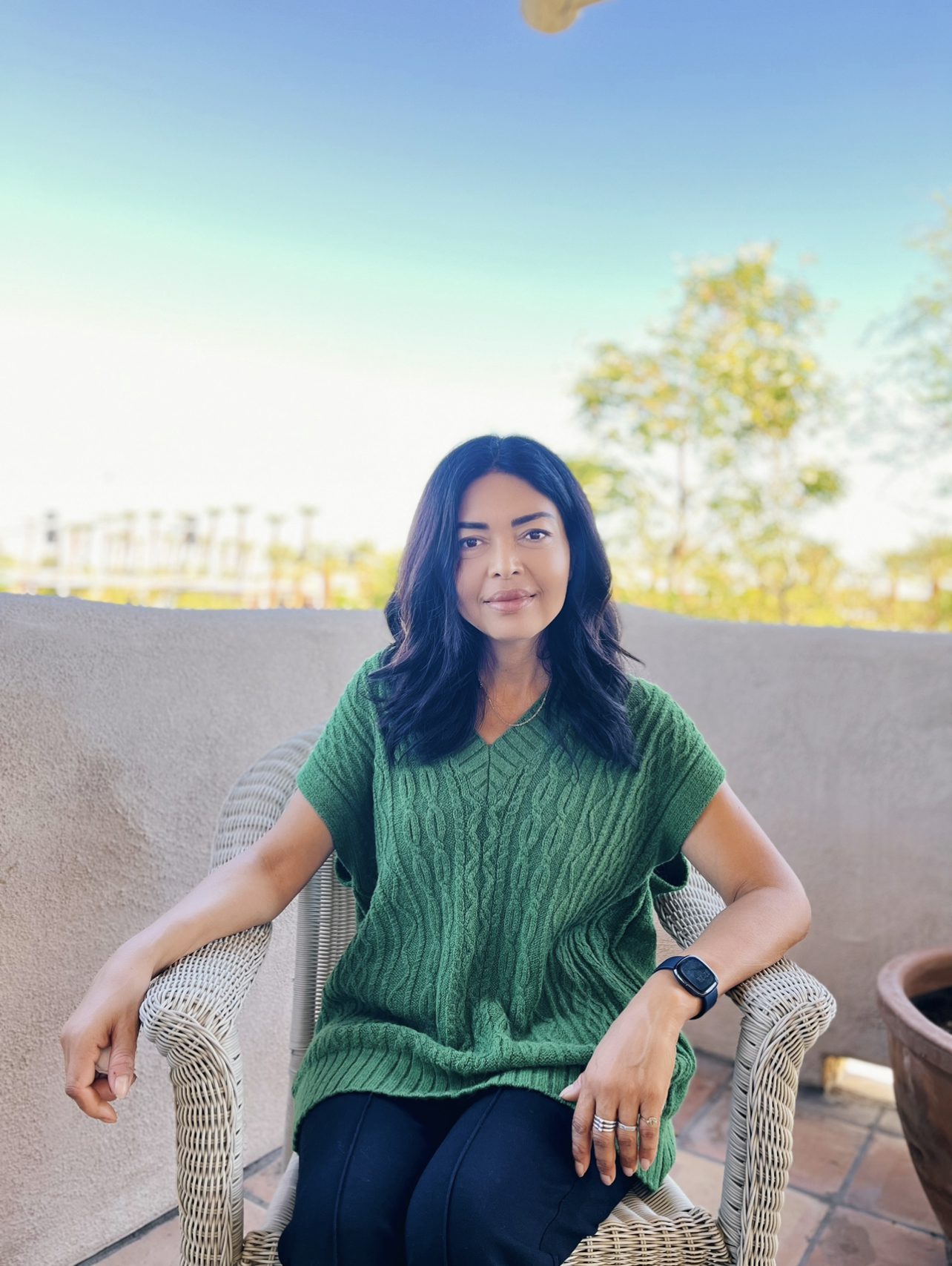 A woman sitting on a wicker chair outdoors wearing a green sweater and black watch, with blurred greenery in the background.